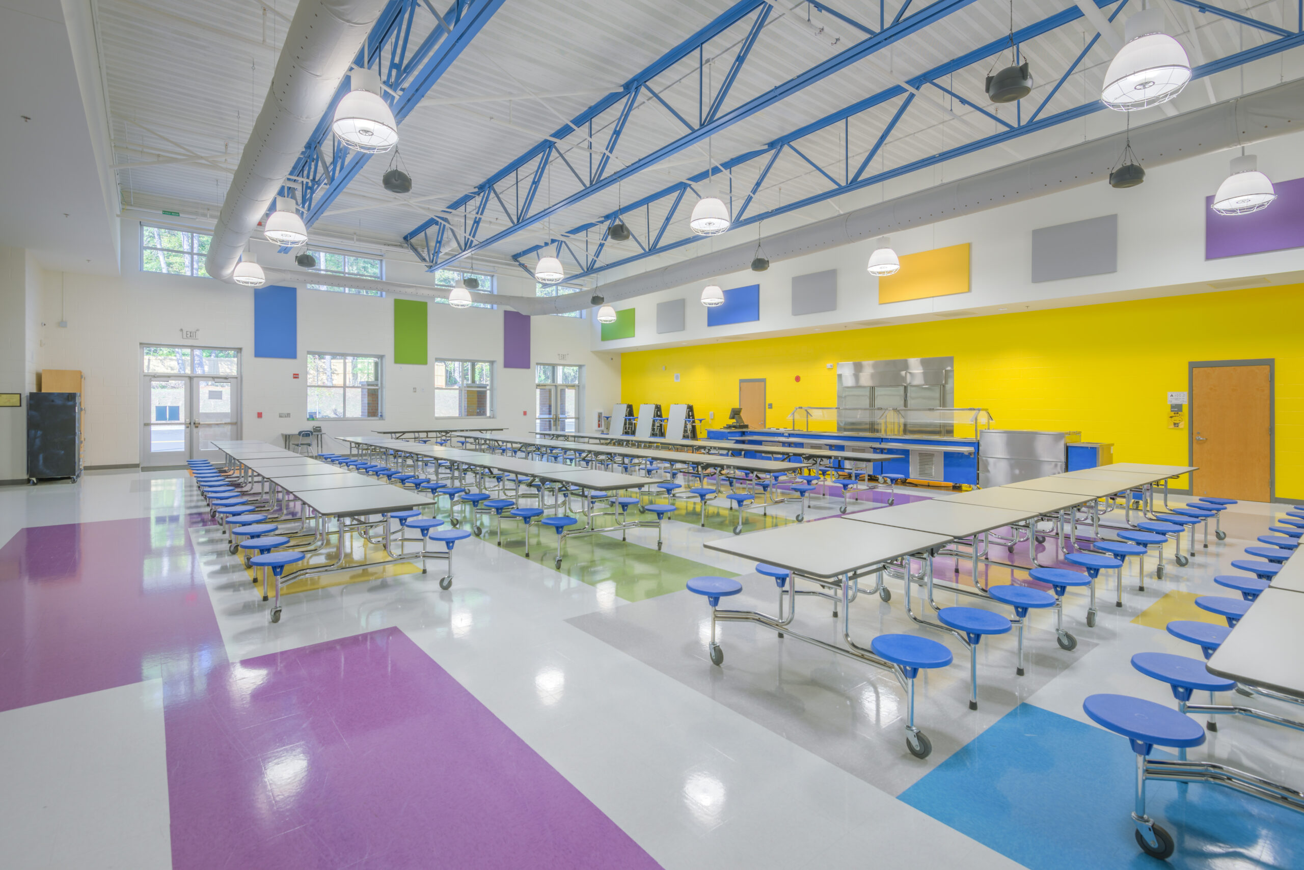Barton Pond Elementary School Cafeteria with Blue Bar Joists, a Yellow Accent Wall, Foldable Lunch Tables with Attached Blue Stools, and a Serving Line